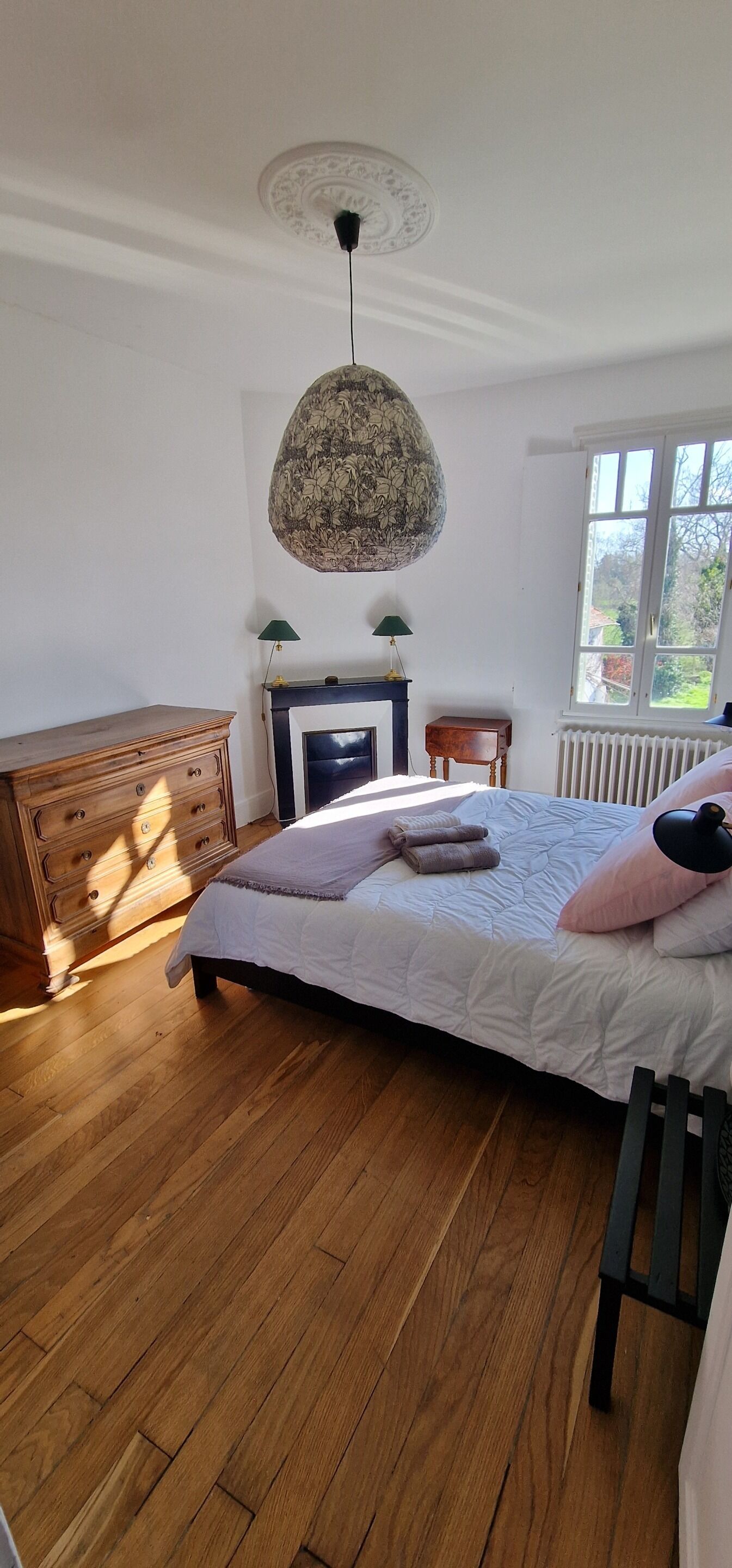 Een slaapkamer met een houten dressoir, een bed met een witte sprei, een hanglamp en een raam met uitzicht.