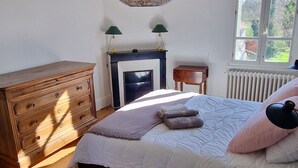 A bedroom with a wooden dresser, a bed with a white quilt, a hanging lamp, and a window with a view.