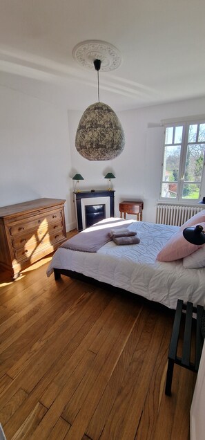 Une chambre à coucher avec une commode en bois, un lit recouvert d’une courtepointe blanche, une lampe suspendue et une fenêtre donnant sur un beau paysage.