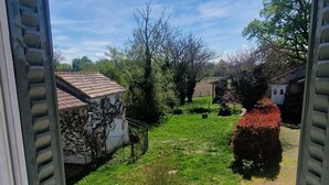 Vista a través de una ventana abierta con un radiador abajo, un césped verde y un edificio blanco con techo rojo al fondo.