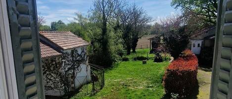 Vista a través de una ventana abierta con un radiador abajo, un césped verde y un edificio blanco con techo rojo al fondo.