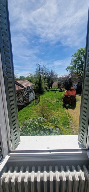 A view through an open window with a radiator below, a green lawn, and a white building with a red roof in the background. - le Pré du Dimanche chambre 3 les Tanneries (Maringues)