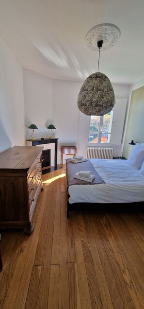 A bedroom with a wooden dresser, a bed with white linens, a hanging lamp, and a window with a view of trees. - le Pré du Dimanche chambre 3 les Tanneries (Maringues)