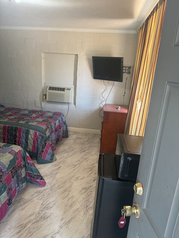 Habitación de hotel con dos camas, televisor montado en la pared, equipo de aire acondicionado, cómoda de madera y puerta con cerradura de llave.