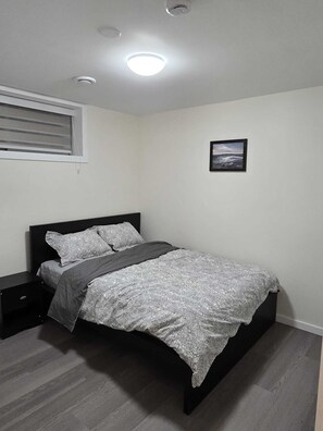 Una camera da letto con un letto, un comodino, una finestra con persiane, una luce a soffitto e un quadro appeso al muro.