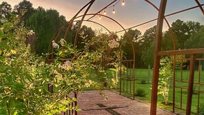 Um caminho de jardim com piso de tijolos, cercado por vegetação e um arco de metal com luzes de cordão.