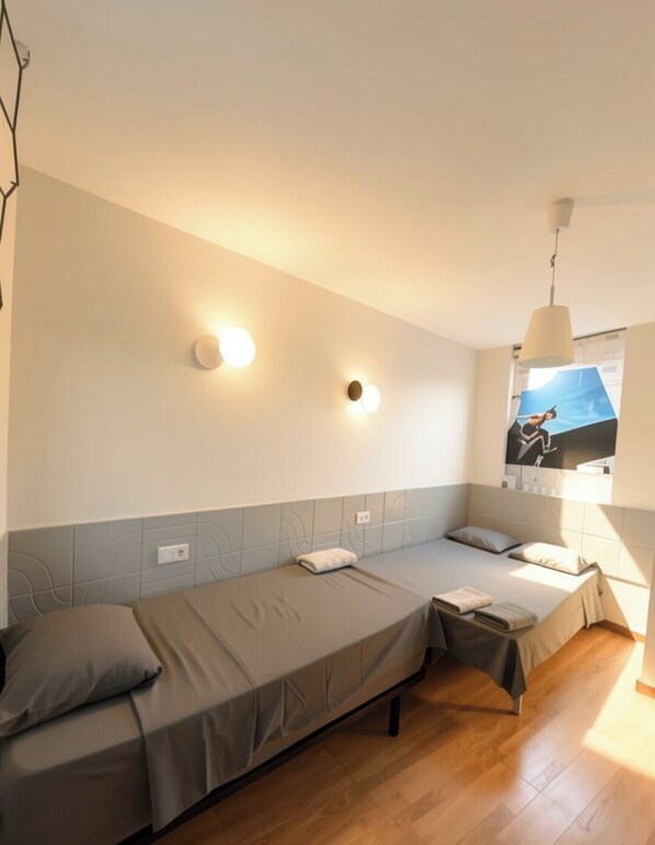 Una stanza con due letti singoli, una lampada fissata al muro, una finestra con un poster e un pavimento in legno.