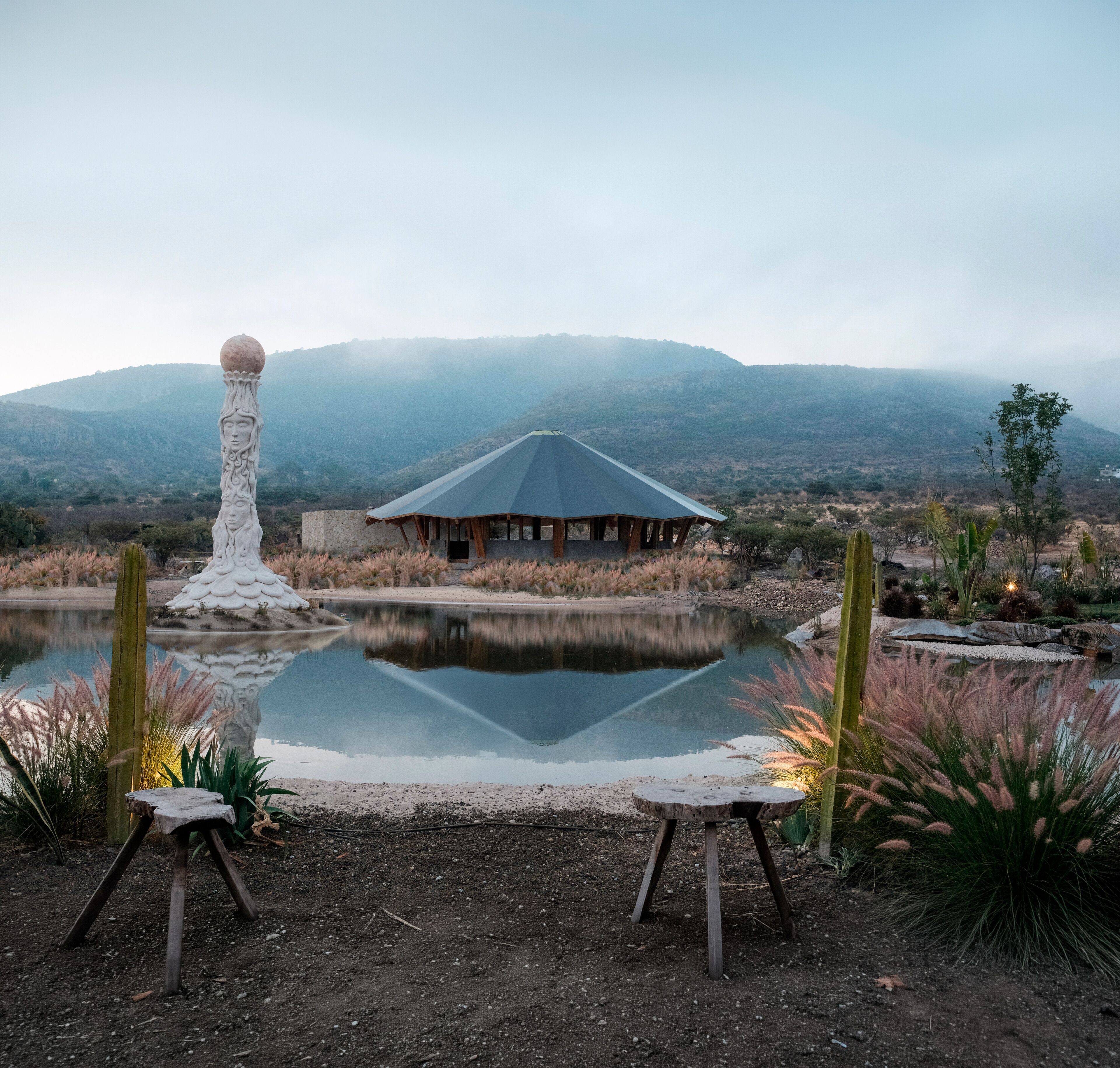 A rustic building with a sloped roof, a pond in the foreground, and a mountainous backdrop.