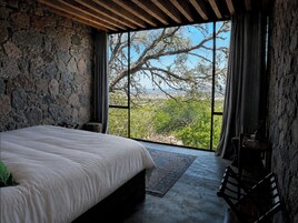 A bedroom with a stone wall, a large bed, a view of trees, and a window with curtains.