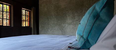 A bed with a white sheet, a teal pillow, and a window with a view of trees.