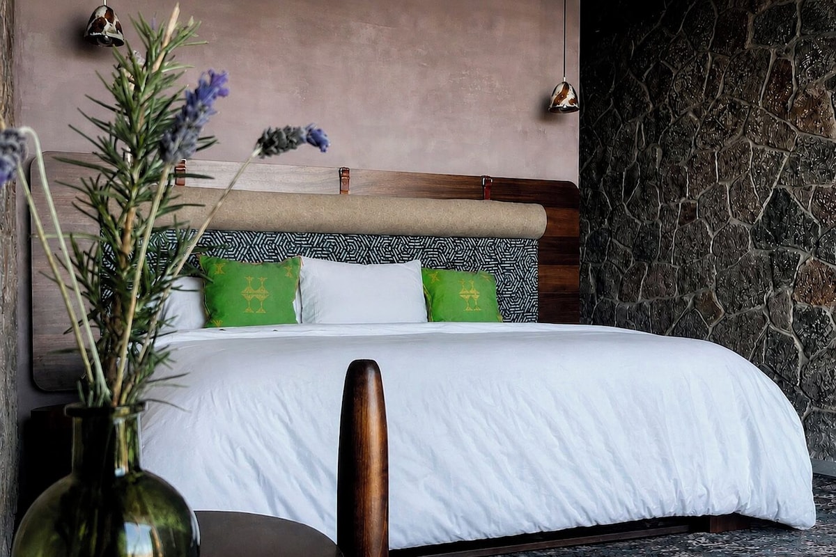 Une chambre à coucher avec un lit, des oreillers, un vase de fleurs et un mur en pierre.
