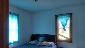 Ein Einzelbett mit gemusterten Kissen, ein Nachttisch, ein Schreibtisch und ein Fenster mit Vorhängen.