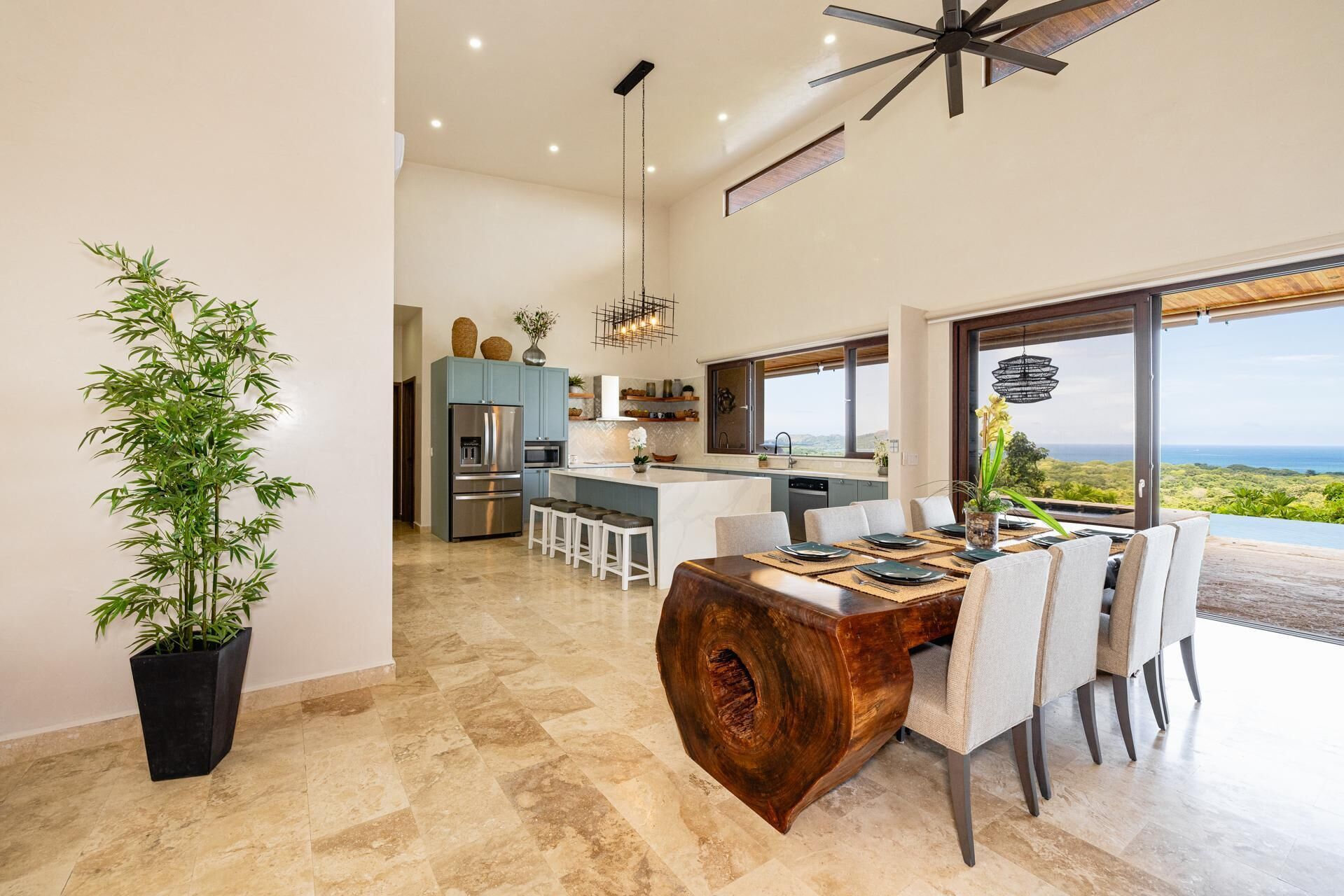 A modern kitchen with a central island, a dining area with a wooden table, and a large window offering a view of the landscape.