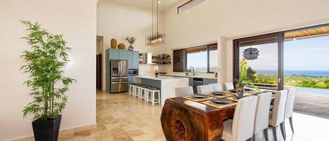 A modern kitchen with a central island, a dining area with a wooden table, and a large window offering a view of the landscape.