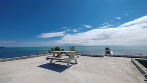 Eine Betonplattform mit einer hölzernen Bank und einem reetgedeckten Gebäude mit Blick auf ein ruhiges Meer und einen klaren Himmel.