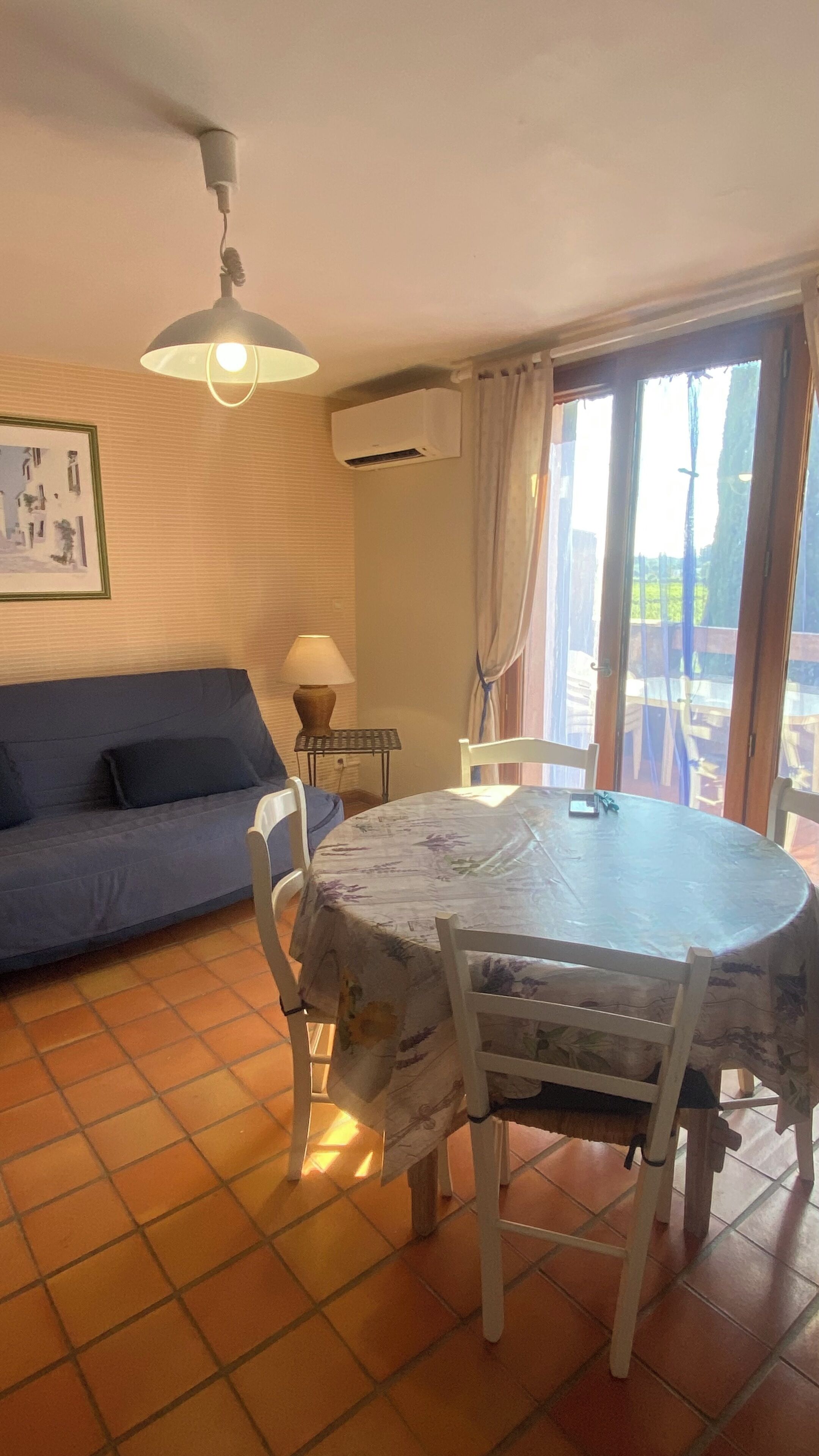 Ein Wohnzimmer mit Sofa, Tisch mit Stühlen, Lampe, Klimaanlage und einem Fenster mit Vorhängen.