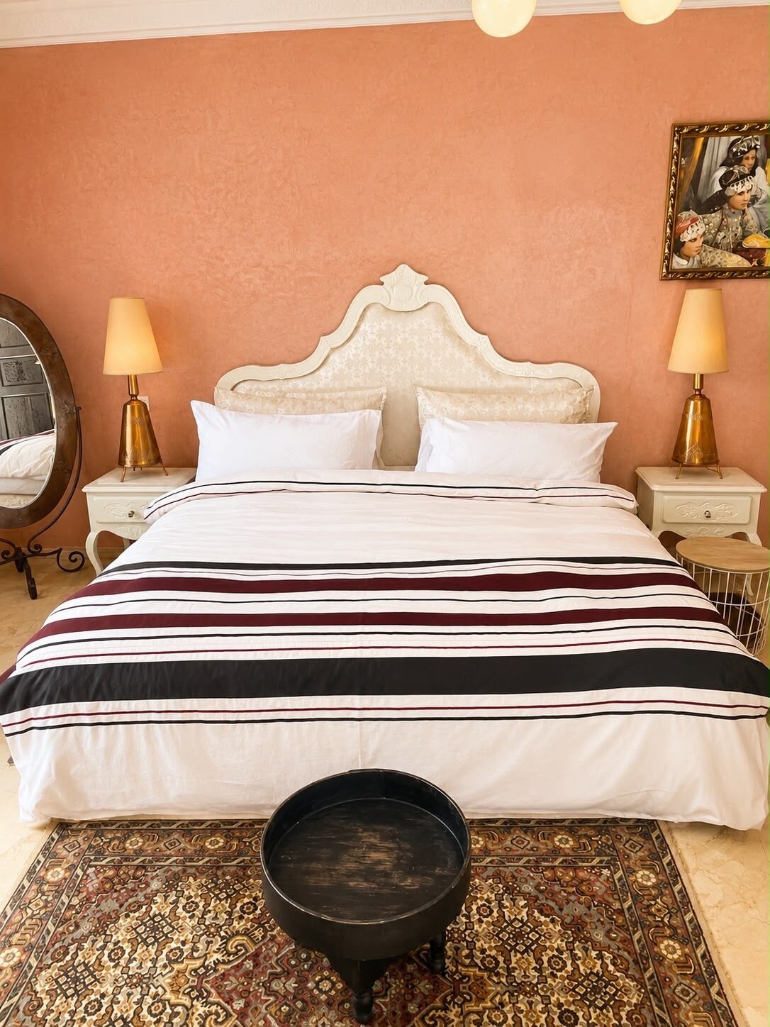 A bed with a striped bedspread, two bedside lamps, a round mirror, a patterned rug, and a small black table.