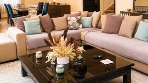 Een woonkamer met een hoekbank, een salontafel met decoratieve voorwerpen, een eethoek met stoelen en een trap.