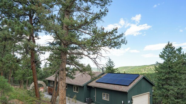 A green building with a garage, solar panels on the roof, and a stone archway. - 3-bdrm home in Evergreen w/sweeping mountain views, hot tub, sauna, & fire place (Evergreen)