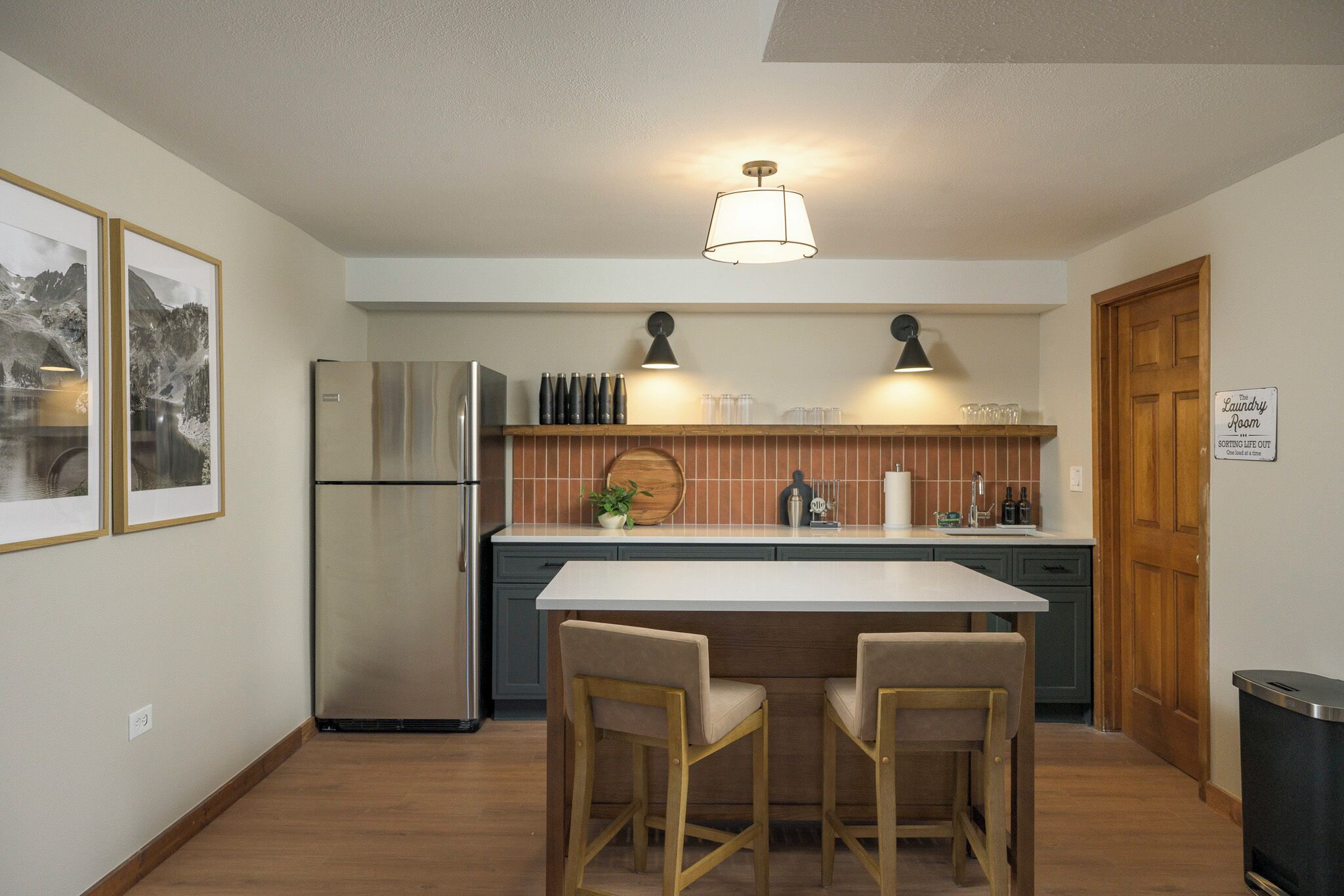 A compact kitchen with a refrigerator, two chairs, a wooden table, and a trash bin.