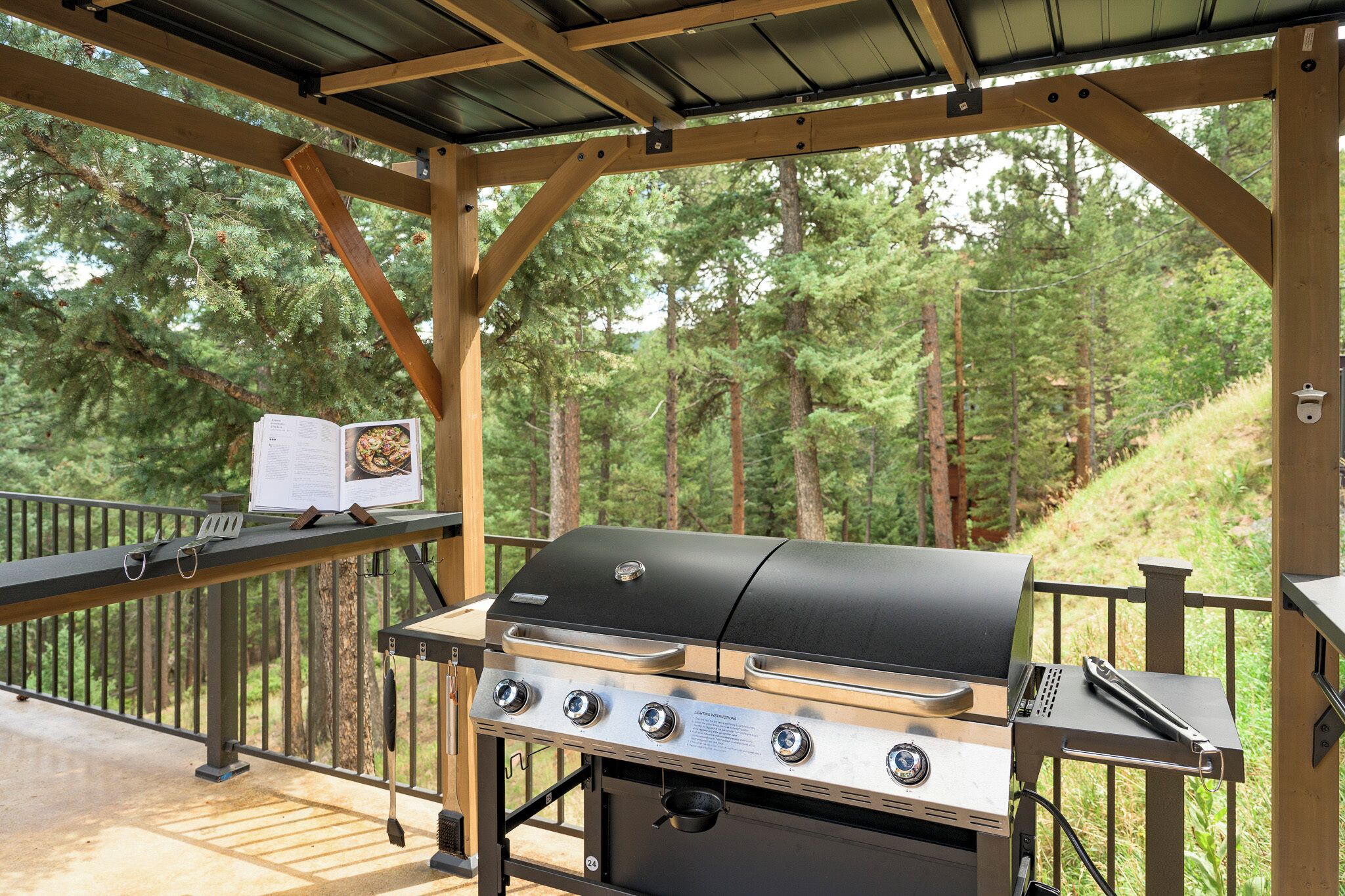 Una estación de parrilla al aire libre con una parrilla metálica, un libro de cocina y un par de pinzas sobre una terraza de madera.