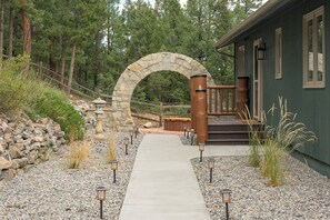 A pathway leading to a stone archway, a wooden deck, and a building with a green exterior. - Luxe 3BD Mountain Retreat w/Hot Tub, Sauna, 98in Theater. 25 mins to Red Rocks (Evergreen)