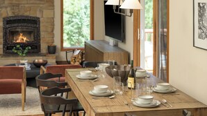 A dining area with a wooden table set for four, surrounded by chairs, with a fireplace and a television in the background. - 3-bdrm home in Evergreen w/sweeping mountain views, hot tub, sauna, & fire place (Evergreen)