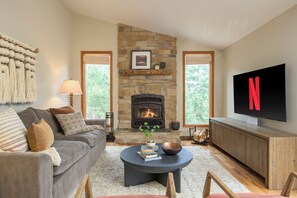 A living room with a stone fireplace, a grey sofa, a coffee table with books, and a TV on a wooden cabinet. - Luxe 3BD Mountain Retreat w/Hot Tub, Sauna, 98in Theater. 25 mins to Red Rocks (Evergreen)