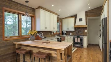 A kitchen with wooden flooring, white cabinets, a central island with stools, and a large window overlooking trees.
