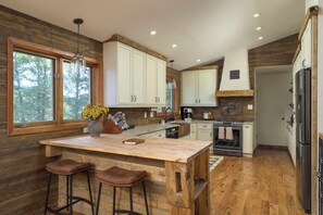 A kitchen with wooden flooring, white cabinets, a central island with stools, and a large window overlooking trees. - Luxe 3BD Mountain Retreat w/Hot Tub, Sauna, 98in Theater. 25 mins to Red Rocks (Evergreen)