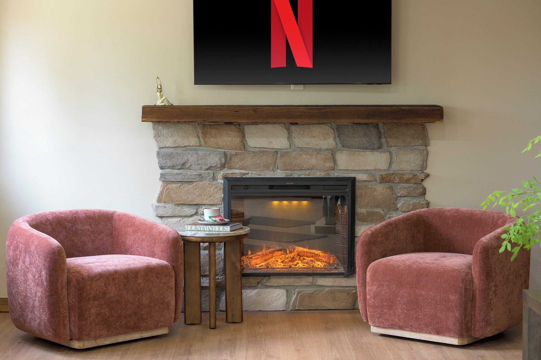 A room with a stone fireplace, two plush armchairs, a small table with a book, and a large TV mounted above the fireplace.