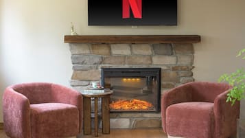 A room with a stone fireplace, two plush armchairs, a small table with a book, and a large TV mounted above the fireplace.