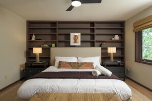 A bedroom with a large bed, two bedside tables, a ceiling fan, and a window with a view of trees.