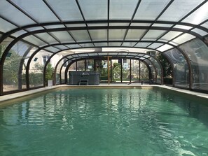 An indoor swimming pool with a glass ceiling, surrounded by a metal frame, and a seating area with a water feature. - 5-star villa with a large pool, hot tub, and sauna in the Loire Valley (Montrichard Val de Cher)