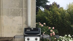 Un barbacoa de gas Weber a l'aire lliure, amb un estenedor negre amb la marca 'Weber' al davant, una planta en test i un contenidor vermell a prop.