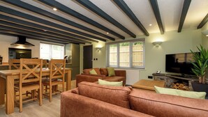A modern living room with a wooden dining table, brown sofas, and a TV. - Two-bedroom cottage for up to four guests, with Wi-Fi, Netflix-ready TV, private decking, Ninja BBQ, hot tub, EV charging and space for up to three dogs. (Cornwall)