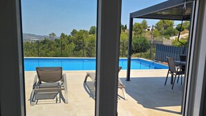 A view through a window of a pool area with lounge chairs and a pergola.