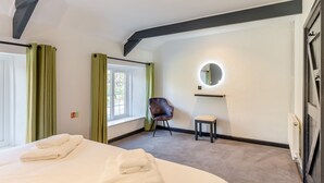 Habitació d'hotel amb un llit, una cadira, un tamboret petit, una finestra amb cortines, un mirall rodó i un radiador.