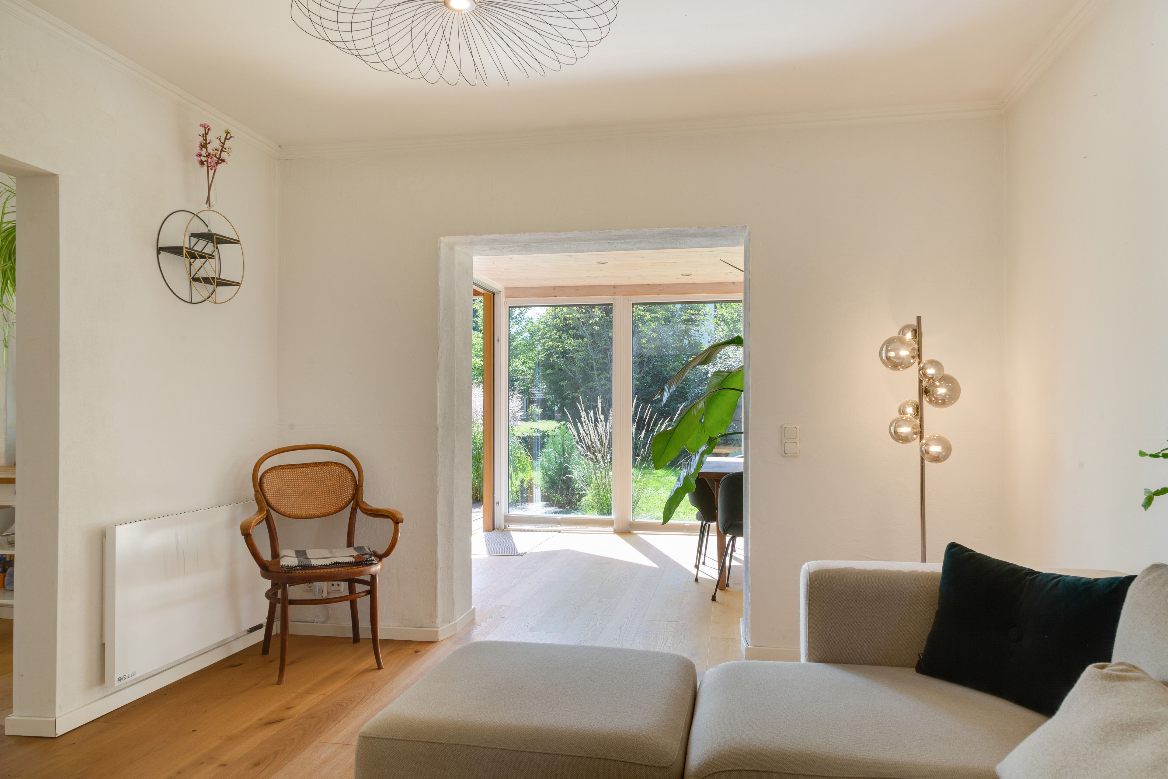 A modern living room with a wooden floor, a sofa, a chair, a floor lamp, and a view of a garden through a glass door.