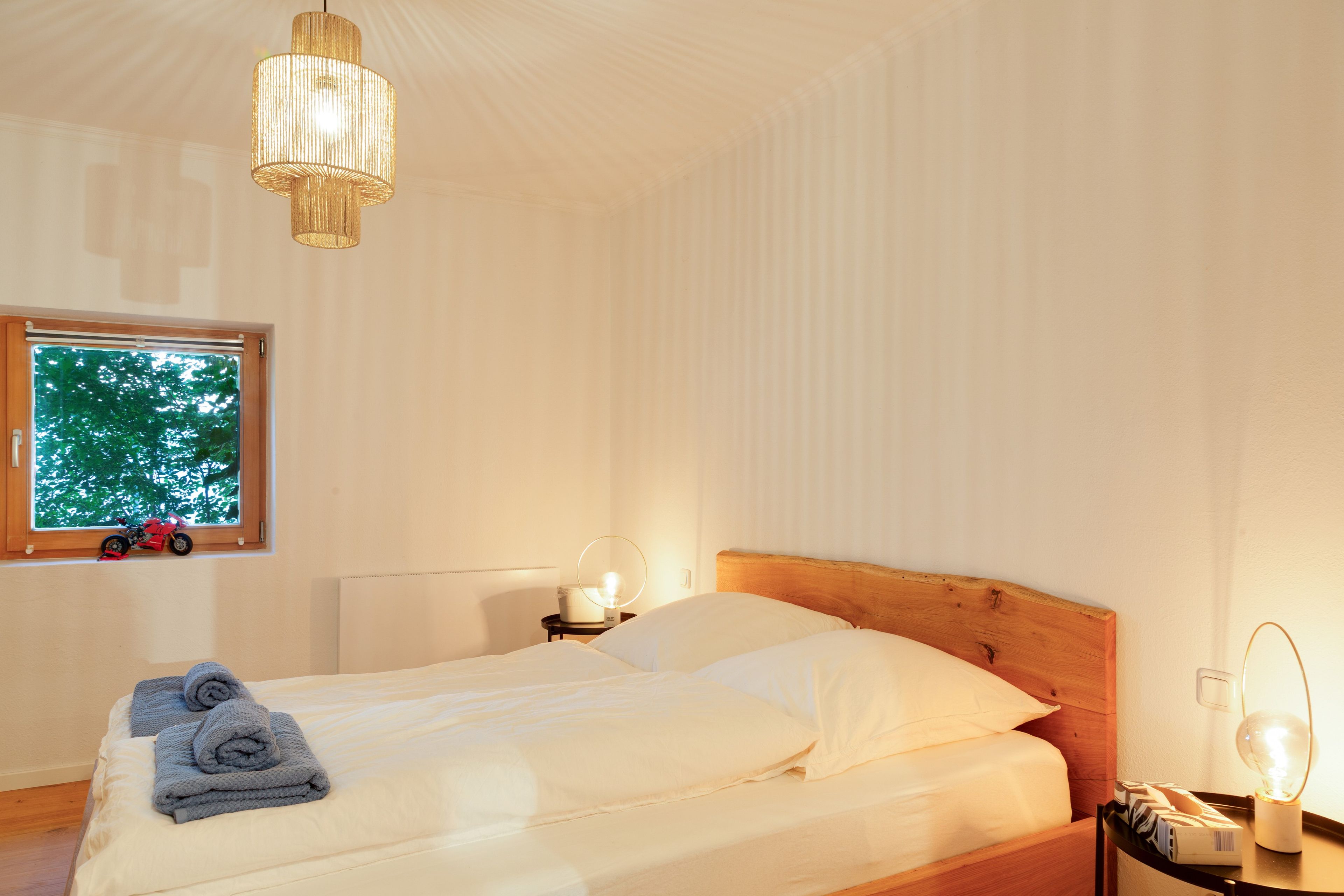 Ein Schlafzimmer mit einem Bett, einem Nachttisch, einer Lampe und einem Fenster mit Blick auf Bäume.