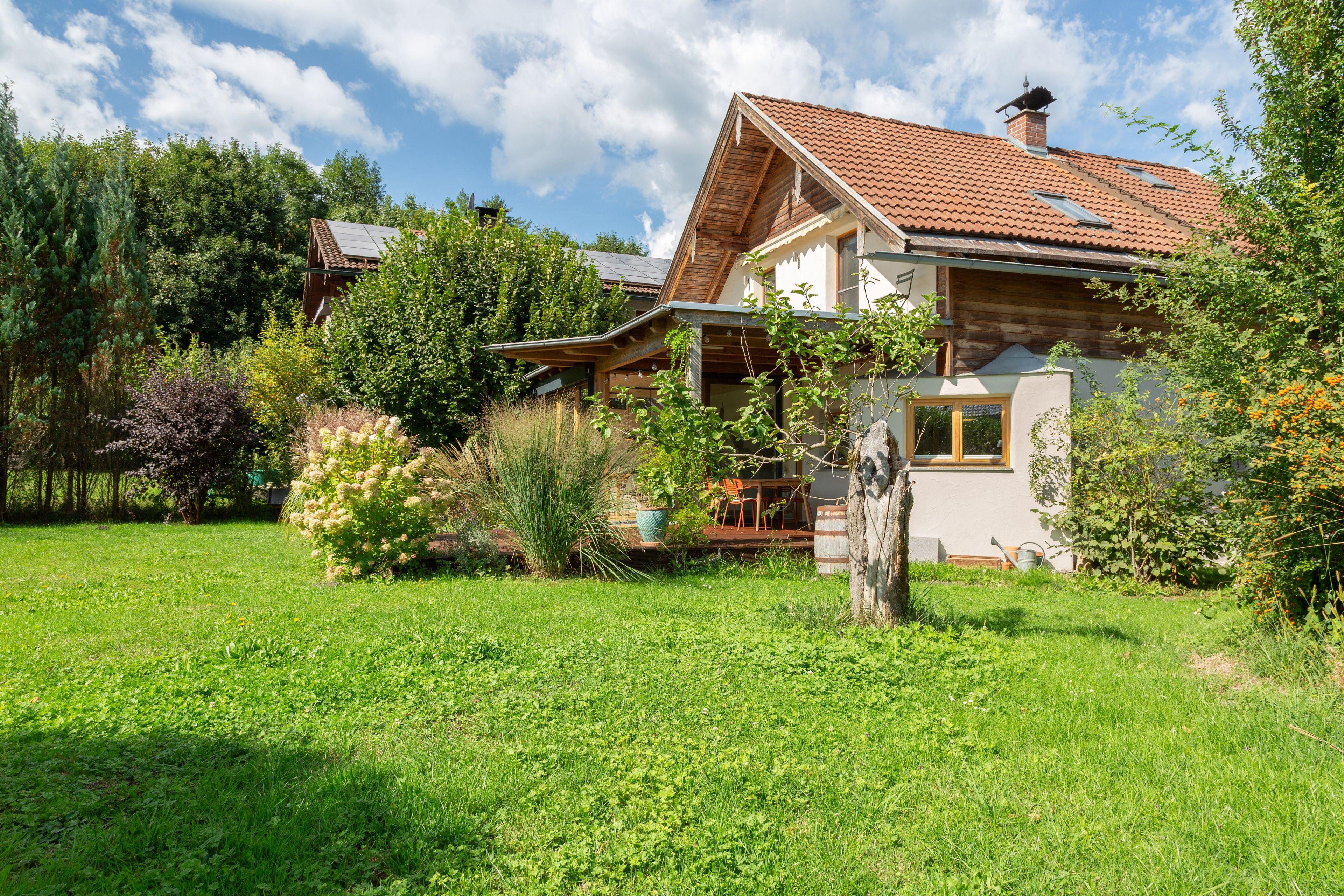 Een huis met een schuin dak, een houten terras en een tuin met verschillende planten en een boom.