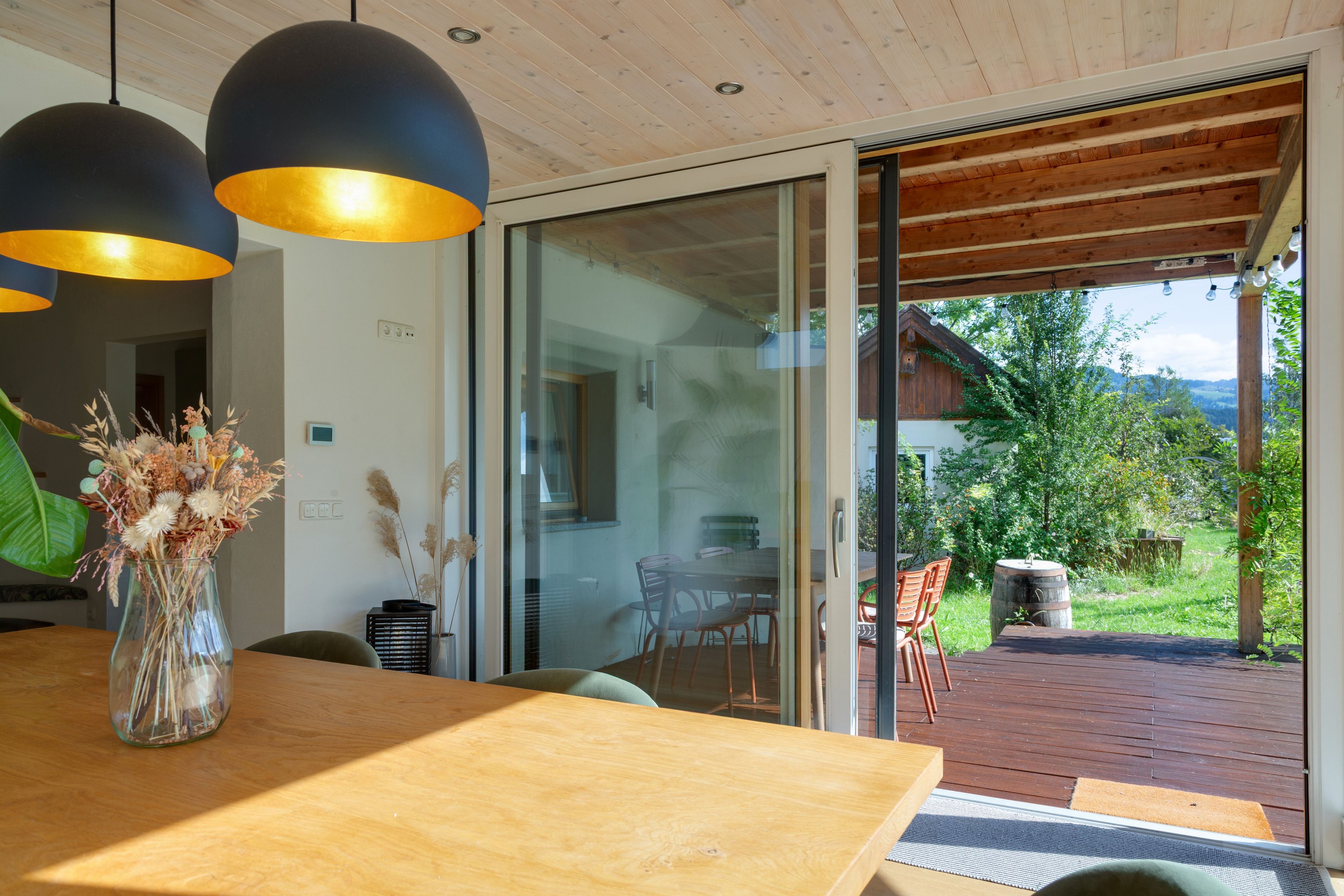 A modern interior with a wooden dining table, a vase with dried flowers, and a view of a wooden deck and greenery outside.