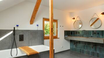 Ein modernes Badezimmer mit einer Badewanne, einer hölzernen Sitzbank, einem Fenster und einem Waschtisch mit Spiegel.