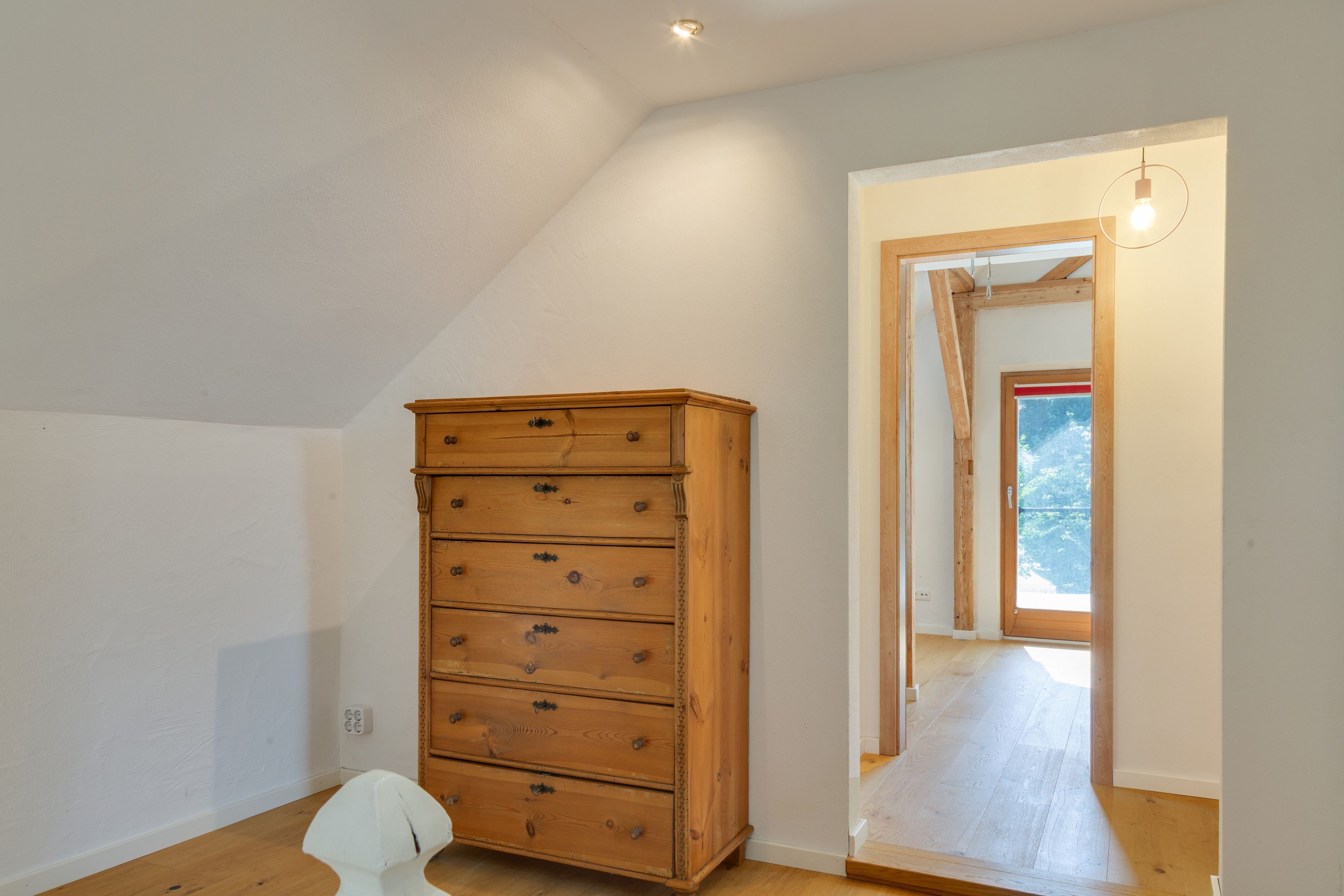 Quarto com cômoda de madeira, uma cadeira branca e porta que dá para uma varanda.