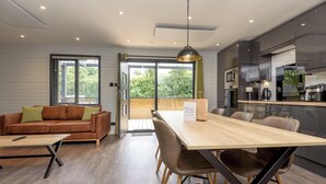 A modern kitchen with a dining area, a sofa, and a large window. - Riverside lodge with spacious decking, Ninja BBQ, Wi-Fi, Netflix-ready TV, EV charging and an outdoor copper bath and shower for a wellness experience in nature. Up to three well-behaved dogs are welcome in selected lodges. (Cornwall)