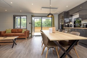 A modern kitchen with a dining area, a sofa, and a large window. - Riverside lodge with spacious decking, Ninja BBQ, Wi-Fi, Netflix-ready TV, EV charging and an outdoor copper bath and shower for a wellness experience in nature. Up to three well-behaved dogs are welcome in selected lodges. (Cornwall)