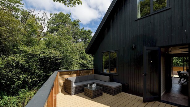 A deck with a built-in sofa and coffee table, surrounded by a wooden deck and a dark building. - Riverside lodge with spacious decking, Ninja BBQ, Wi-Fi, Netflix-ready TV, EV charging and an outdoor copper bath and shower for a wellness experience in nature. Up to three well-behaved dogs are welcome in selected lodges. (Cornwall)