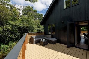 A deck with a built-in sofa and coffee table, surrounded by a wooden deck and a dark building. - Riverside lodge with spacious decking, Ninja BBQ, Wi-Fi, Netflix-ready TV, EV charging and an outdoor copper bath and shower for a wellness experience in nature. Up to three well-behaved dogs are welcome in selected lodges. (Cornwall)