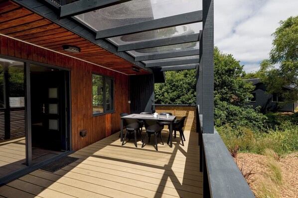 A modern outdoor deck with a wooden floor, a table with chairs, and a glass enclosure. - Riverside lodge with spacious decking, Ninja BBQ, Wi-Fi, Netflix-ready TV, EV charging and an outdoor copper bath and shower for a wellness experience in nature. Up to three well-behaved dogs are welcome in selected lodges. (Cornwall)
