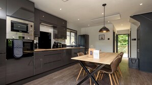 A modern kitchen with dark cabinetry, a built-in microwave, and a wooden dining table with chairs. - Riverside lodge with spacious decking, Ninja BBQ, Wi-Fi, Netflix-ready TV, EV charging and an outdoor copper bath and shower for a wellness experience in nature. Up to three well-behaved dogs are welcome in selected lodges. (Cornwall)